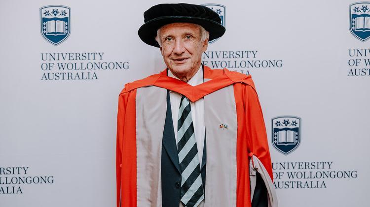 Professor Colin D Woodroffe in orange Honorary Gown in front of a white wall with repeated UOW logo