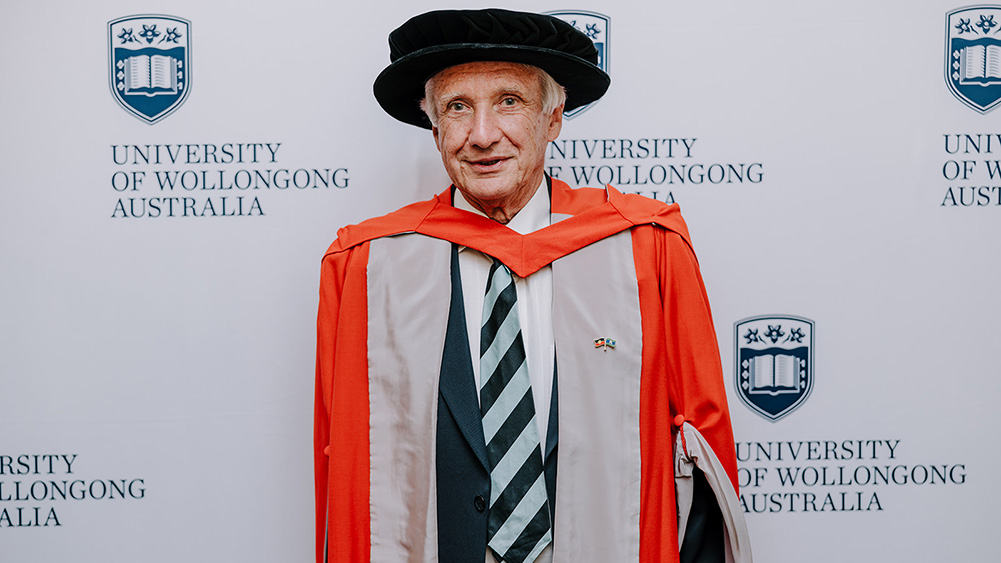 Professor Colin D Woodroffe in orange Honorary Gown in front of a white wall with repeated UOW logo