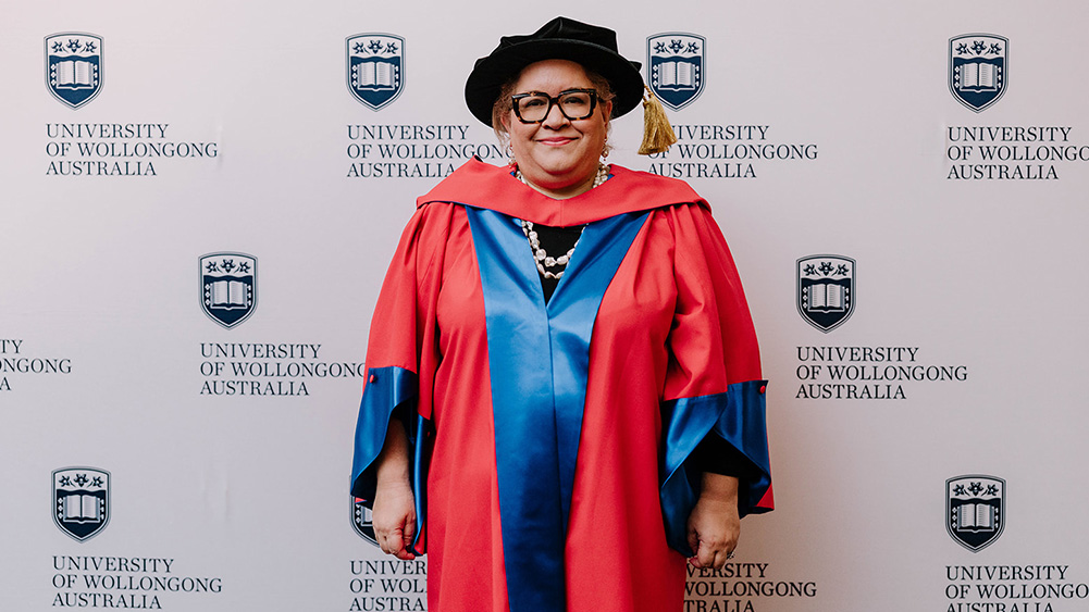 Professor Megan Davis AC in red Honorary Gown in front of a white wall with repeated UOW logo