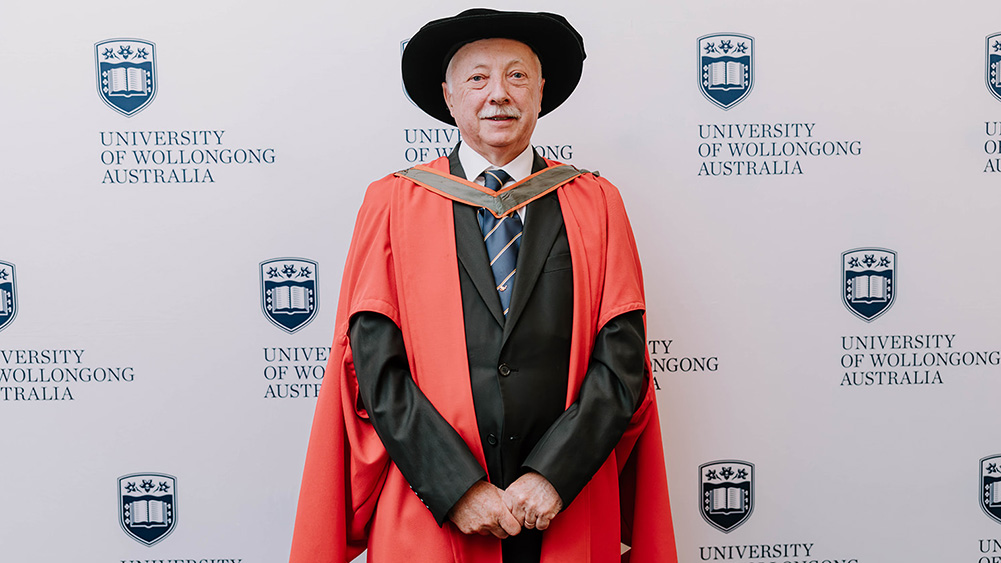 Professor Timothy John McCarthy in red Honorary Gown in front of a white wall with repeated UOW logo