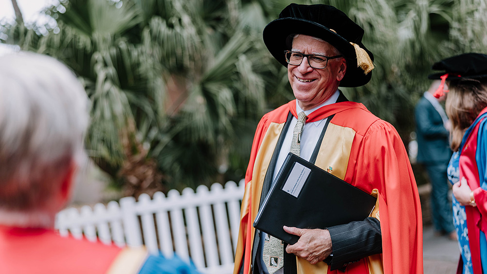 Senior Professor Colin Murray-Wallace wears a black cap and glasses, holds a folder. He stands outdoors with palm trees in the background, conversing with others.