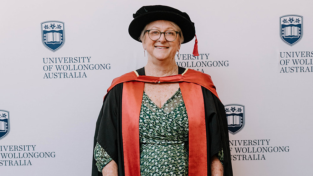 Senior Professor Kristine French in a black Honorary Gown in front of a white wall with repeated UOW logo