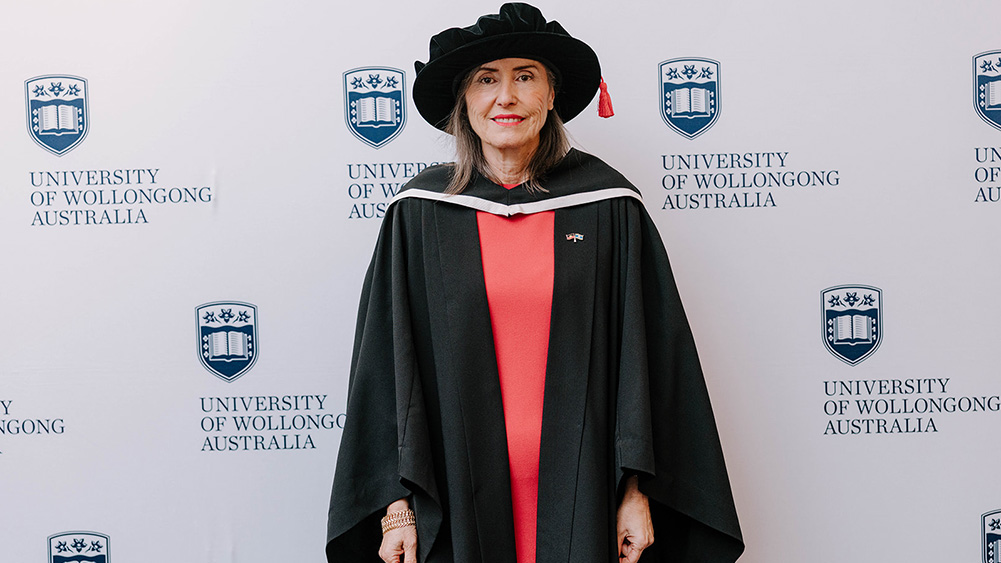 Senior Professor Marie Ranson in a black Honorary Gown in front of a white wall with repeated UOW logo