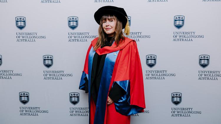Tara June Winch in red Honorary Gown in front of a white wall with repeated UOW logo