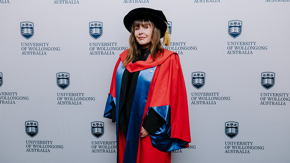Tara June Winch in red Honorary Gown in front of a white wall with repeated UOW logo
