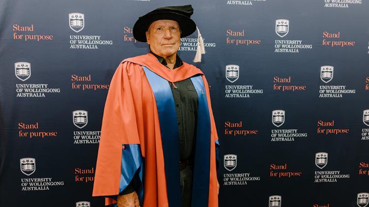 William Buttemer in Honorary Gown in front of navy wall with repeated UOW logo