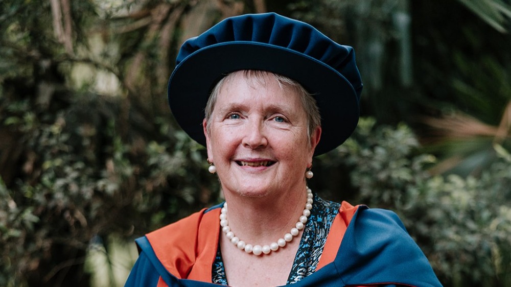 A portrait of Grace McCarthy, wearing a blue pleated academic Tudor bonnet and blue and red regalia. She is wearing a pearl necklace and matching pearl drop earrings, standing outdoors with a soft-focus background of green foliage.