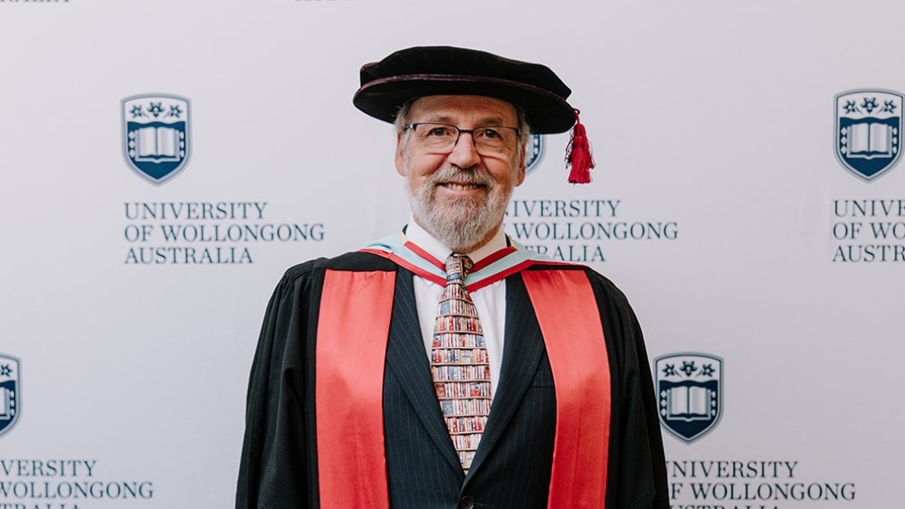 Peter McLennan in black and red academic regalia and a patterned bookshelf tie, posing in front of a University of Wollongong branded backdrop.