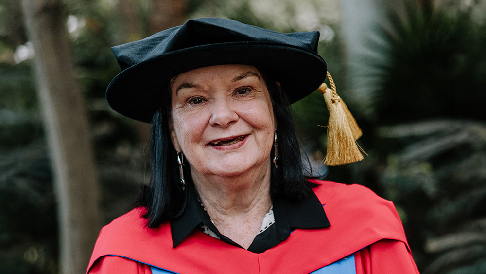 A close-up portrait of Sharan Burrow. She is wearing red and blue academic regalia and a black velvet mortarboard with a gold tassel. She has dark hair and is wearing silver drop earrings, standing against a soft-focus outdoor background of greenery.