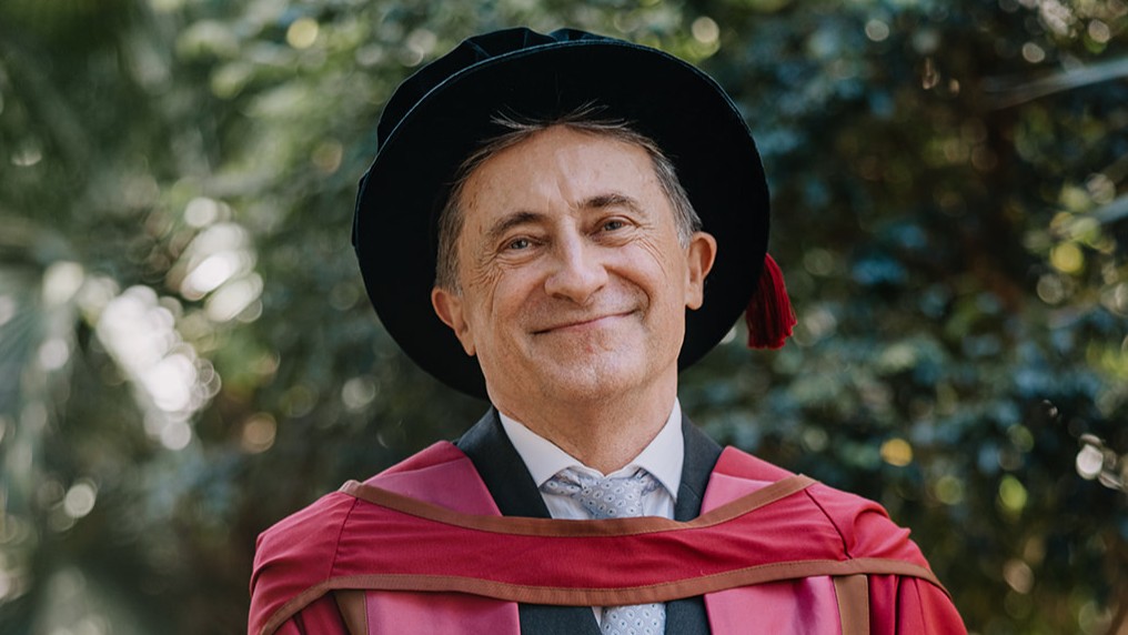 Close-up portrait of Simon Ville, wearing deep red and pink academic regalia with a black velvet Tudor bonnet featuring a red tassel. He is wearing a white shirt and a patterned silver tie, standing outdoors against a blurred background of green trees and dappled sunlight.