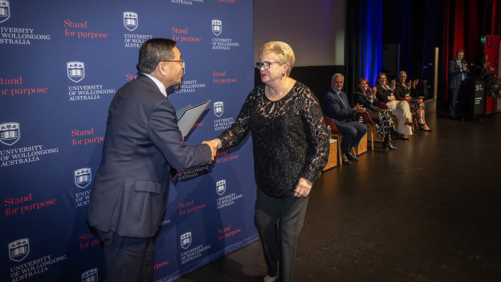Kerrie Gamble receives recognition at the University of Wollongong Vice-Chancellor’s Awards ceremony, shaking hands with UOW Vice-Chancellor and President Max Lu on stage. Other attendees are seated in the background, with University of Wollongong branding and “Stand for Purpose” messaging displayed on a backdrop behind them.