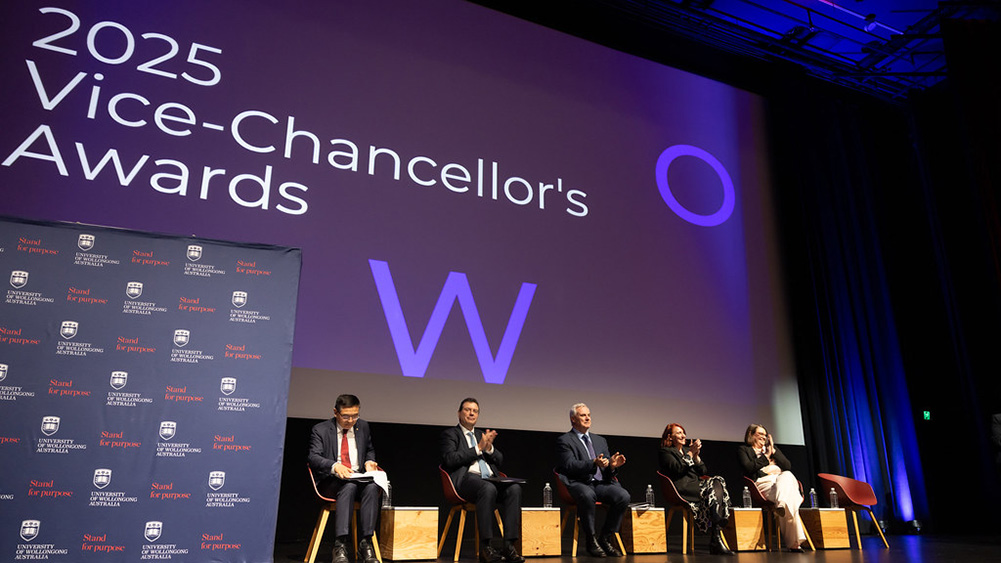 A wide view of an auditorium stage during the 2025 Vice-Chancellor’s Awards event. Large white text reading “2025 Vice-Chancellor’s Awards” is projected on a screen above the stage, with a large purple “W” logo and circular graphic. Five seated panelists are arranged across the stage on chairs, with water bottles placed nearby. A University of Wollongong branded backdrop stands to the left