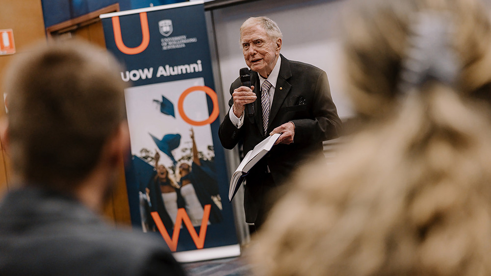 Justice Michael Kirby speaks into a handheld microphone during a student seminar, holding an open book while addressing an audience. A UOW Alumni banner stands behind the speaker, and audience members are visible in the foreground of the indoor event space.