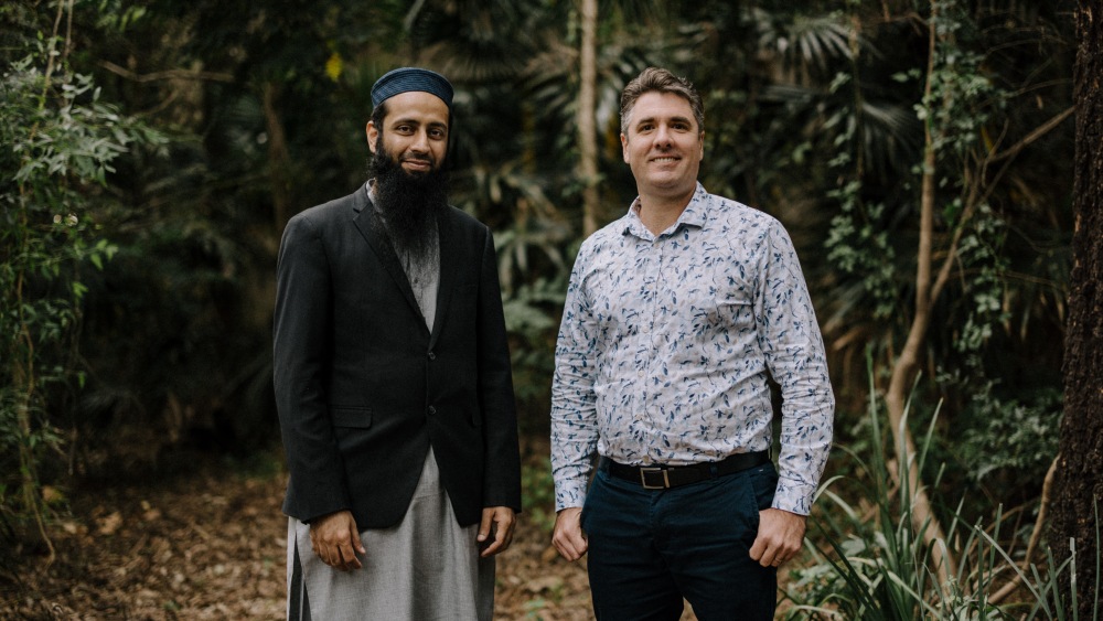 Two men stand in a lush forest setting, one wearing a jacket suit and skullcap, the other in a patterned shirt. Both smile, conveying a friendly tone.