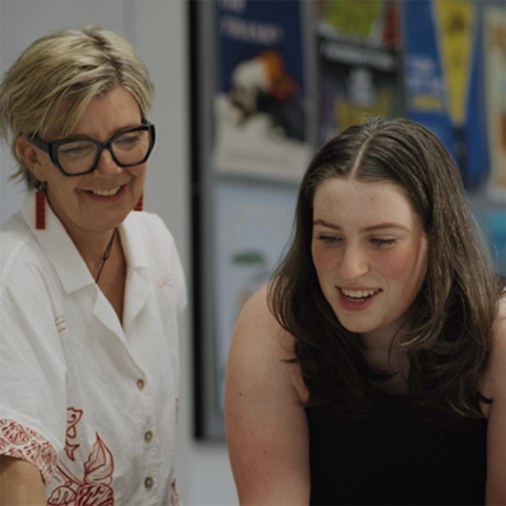 Lecturer Jo Stirling works alongside a student in a classroom setting, reviewing material together at a table. Both are focused on the task, with educational posters displayed on the wall in the background, highlighting a collaborative teaching and learning environment.