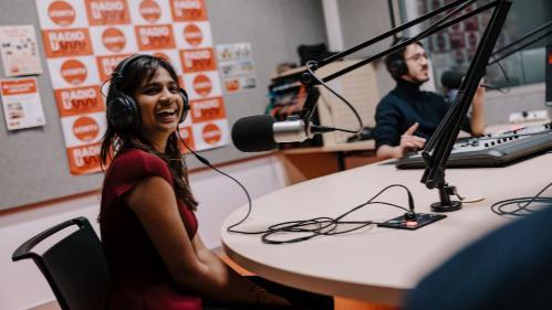 Young woman with headphones smiles at a radio studio desk, a man operates equipment in the background. The atmosphere is lively and engaging.
