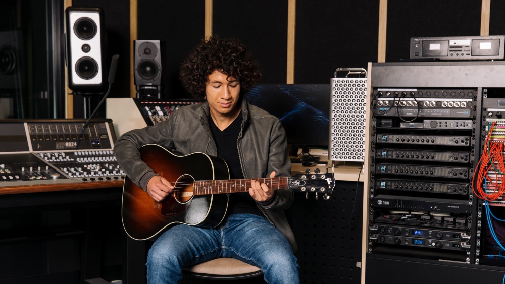 A musician with curly hair sits in a recording studio, playing an acoustic guitar. Surrounding equipment includes speakers, mixing consoles, and cables, creating a focused and creative atmosphere.