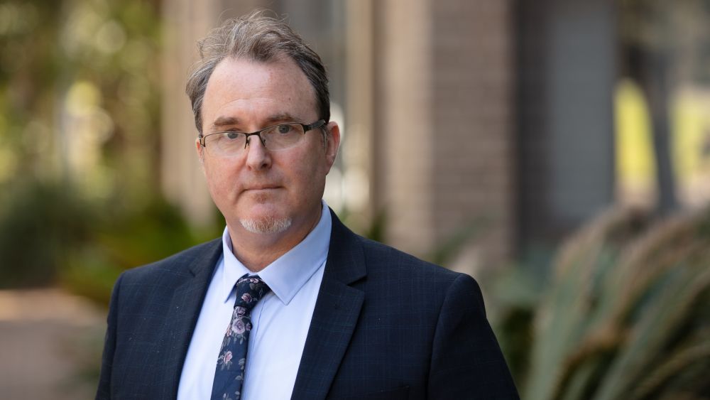 A man with glasses, wearing a suit and blue shirt, looks directly at the camera. A blurred green and beige background conveys a calm tone.