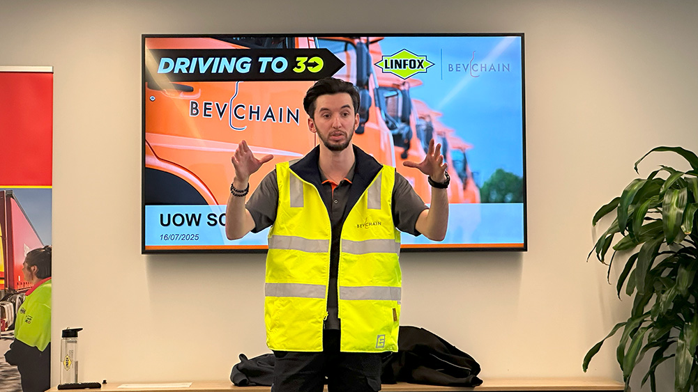 A speaker in a yellow safety vest gestures during a presentation, with Linfox branded graphics in the background.