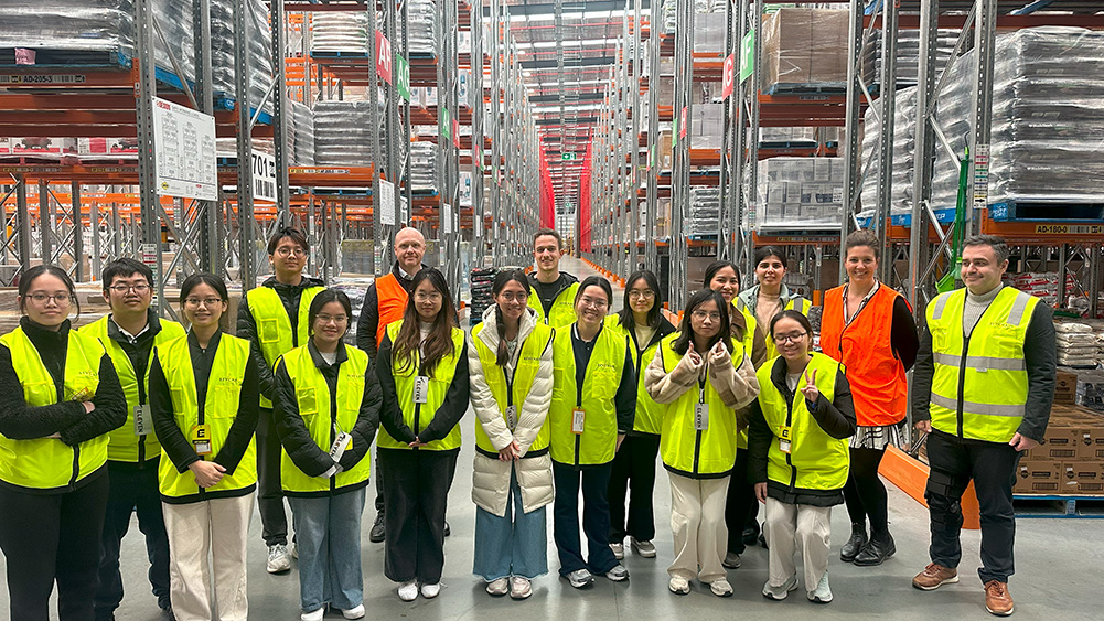 A group of people in bright yellow safety vests poses in a warehouse aisle lined with shelves stacked with goods.