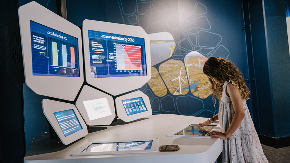 Child interacting with a touchscreen exhibit at a science and sustainability display. Multiple digital screens show graphs, charts and environmental data, including information about emissions reduction targets and data-driven decision-making. A large wall graphic featuring renewable energy imagery, including wind turbines, forms the backdrop to the interactive learning space.