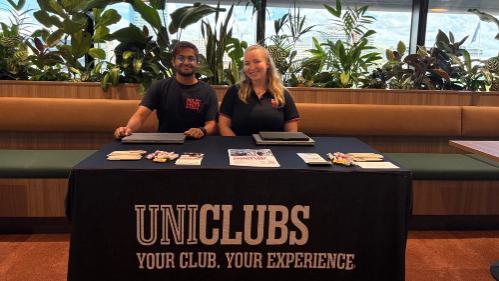 Two staff members sit behind a table displaying promotional materials and a banner for uni clubs, surrounded by greenery.
