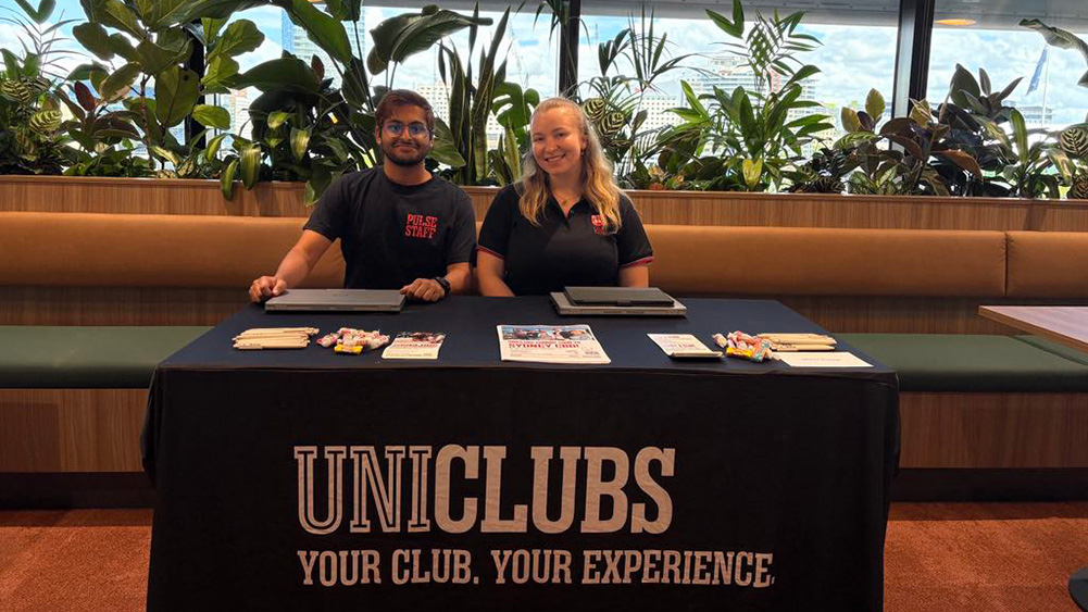 Two staff members sit behind a table displaying promotional materials and a banner for uni clubs, surrounded by greenery.