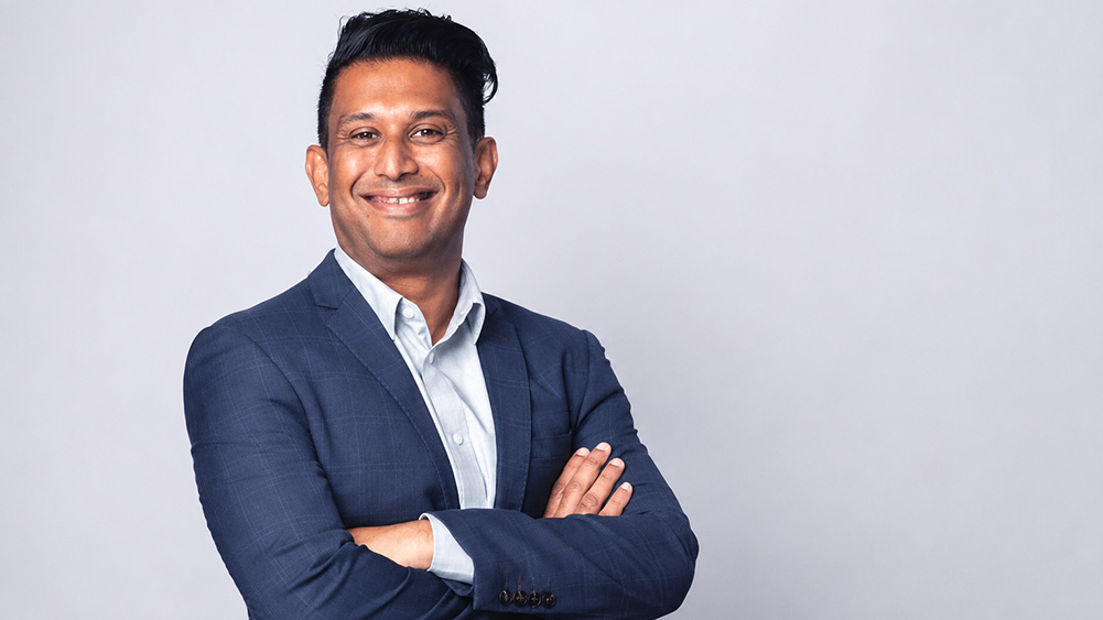 Giri Shanker, Manager, at Sydney CBD Campus wears  a blue suit jacket and light shirt stands smiling with arms crossed against a plain light background.
