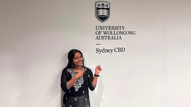 Sydney Business School, UOW student, Gopika Radha smiles as she points to the Sydney Business School, UOW logo on a white wall behind her.