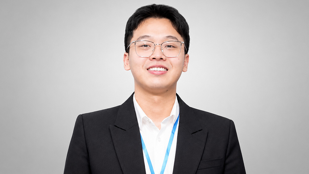 Huu Kim Lam Le wearing a black blazer and white shirt, with a blue lanyard, standing against a light grey background.