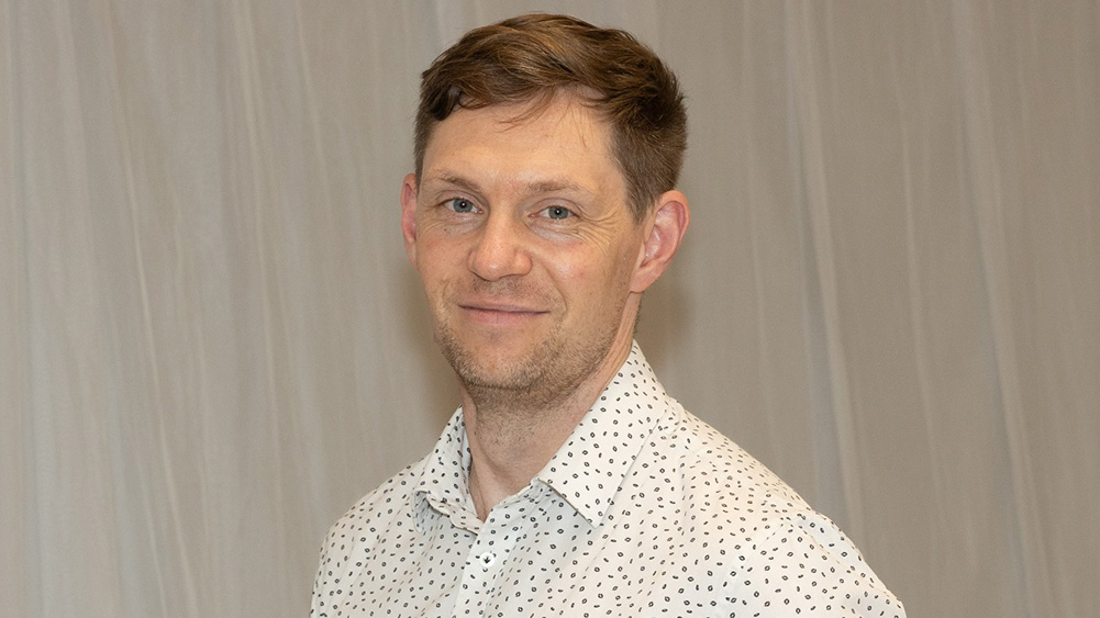 Jackson Howard, Sydney CBD Campus Manager, with short hair smiles softly at the camera, wearing a white shirt with a dotted pattern. The background is a plain, light-colored wall.