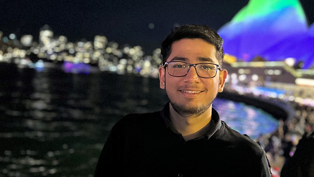 Sydney Business School, UOW Alumni Mahammad Kadwal stands near water at night, with a vibrant city skyline and illuminated buildings reflecting on the water
