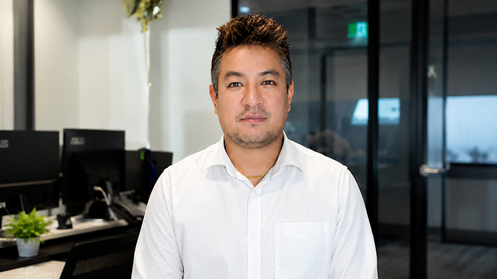 Puran Pradhan, Campus Service Officer at the Sydney CBD Campus, standing in an office workspace wearing a white button-up shirt. Computer monitors, office furniture and glass-walled meeting rooms are visible in the background.
