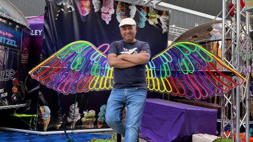 Tomas Sanchez stands confidently in front of colourful neon wings at a vibrant food festival backdrop, showcasing an ice cream theme.