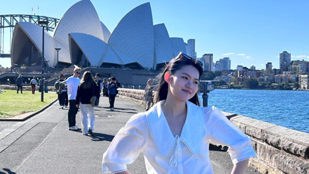 A person in a white blouse poses happily near Sydney Opera House with blue sky and ocean in the background. Others stroll nearby, creating a lively atmosphere.