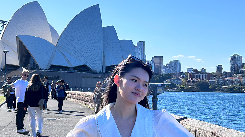 A person in a white blouse poses happily near Sydney Opera House with blue sky and ocean in the background. Others stroll nearby, creating a lively atmosphere.