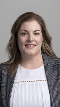 Portrait of Lauren Wise. Lauren wears a white blouse with a Grey jacket, she is in the foreground of a grey wall