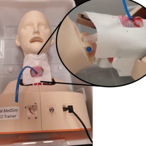 Close-up of a medical simulation airway training mannequin connected to tubing and control equipment, with an inset image highlighting a simulated surgical or intubation procedure on the neck area.