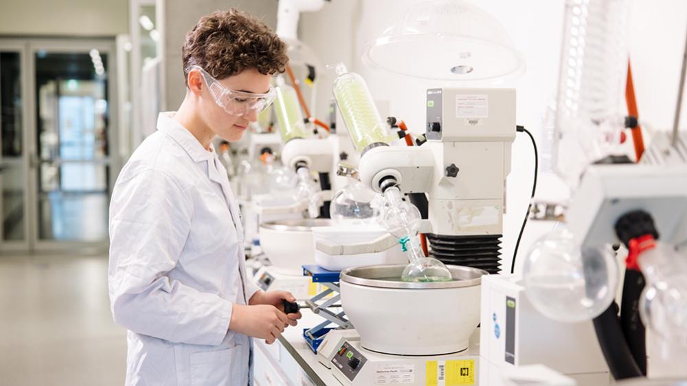 A young scientist in a lab coat operates equipment in a modern laboratory filled with glassware and scientific tools.