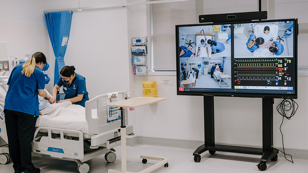 Two healthcare workers in blue uniforms provide patient care beside a hospital bed, with a large screen displaying live monitoring data and images.