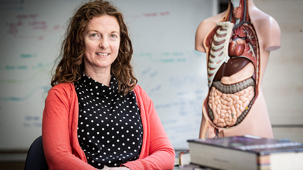 A woman in a polka dot blouse and coral cardigan sits beside a detailed human anatomy model, with educational notes in the background.