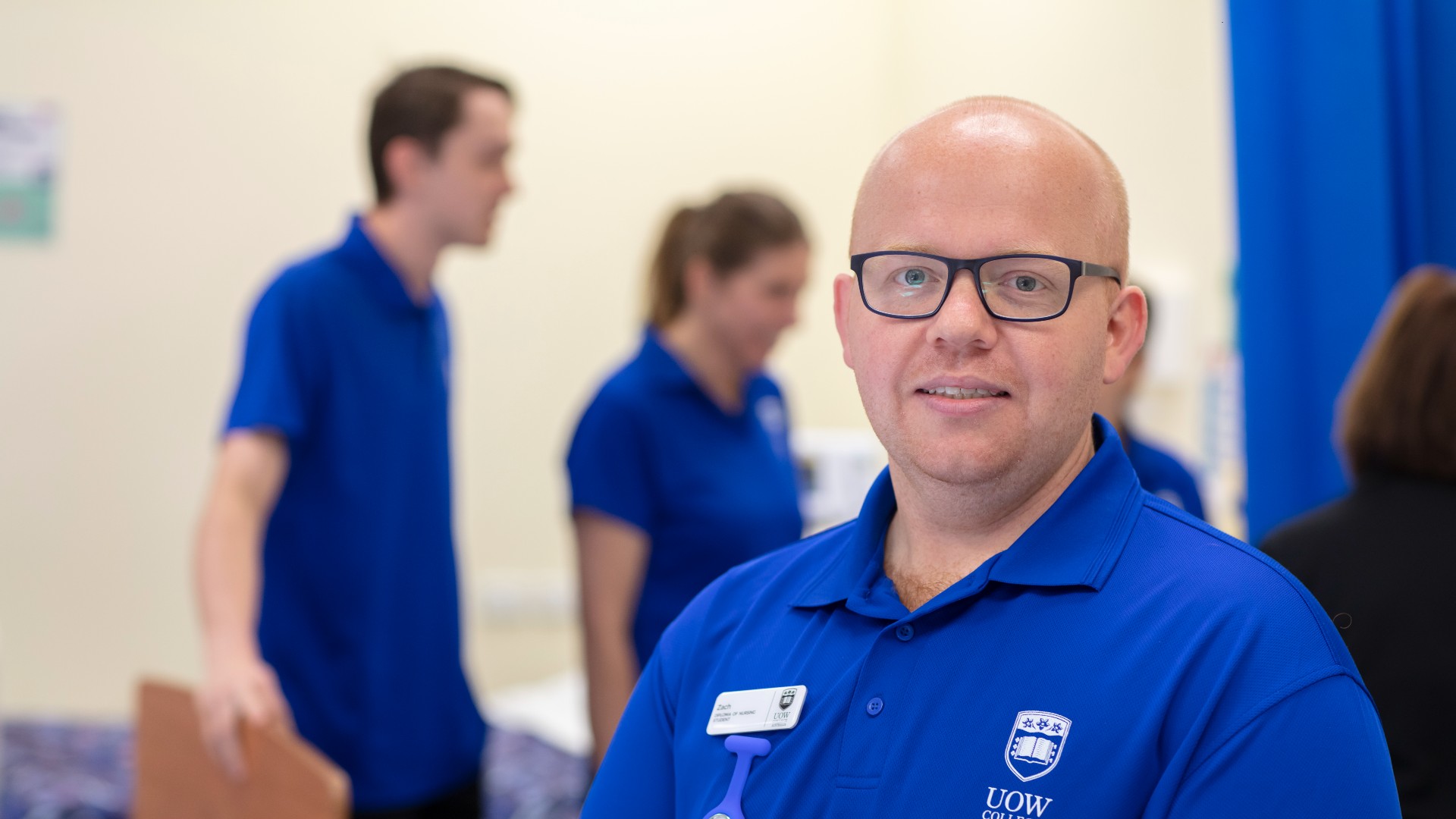 UOW College Nursing student, Zack Pettit wearing blue polo shirt uniform with other nursing students practicing in backgrund.