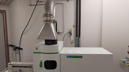 A laboratory setup featuring a scientific instrument with a metal vent, surrounded by pipes and control knobs. The room is sterile and industrial./