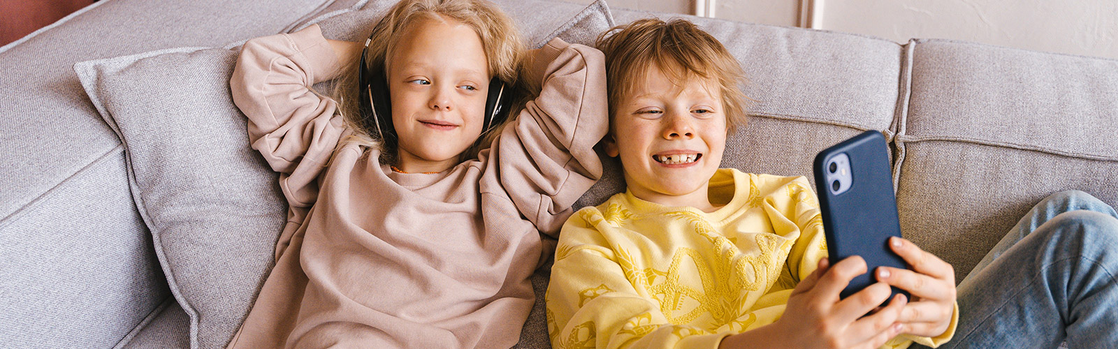 2 kids laying on the couch with headphones on looking at a digital device