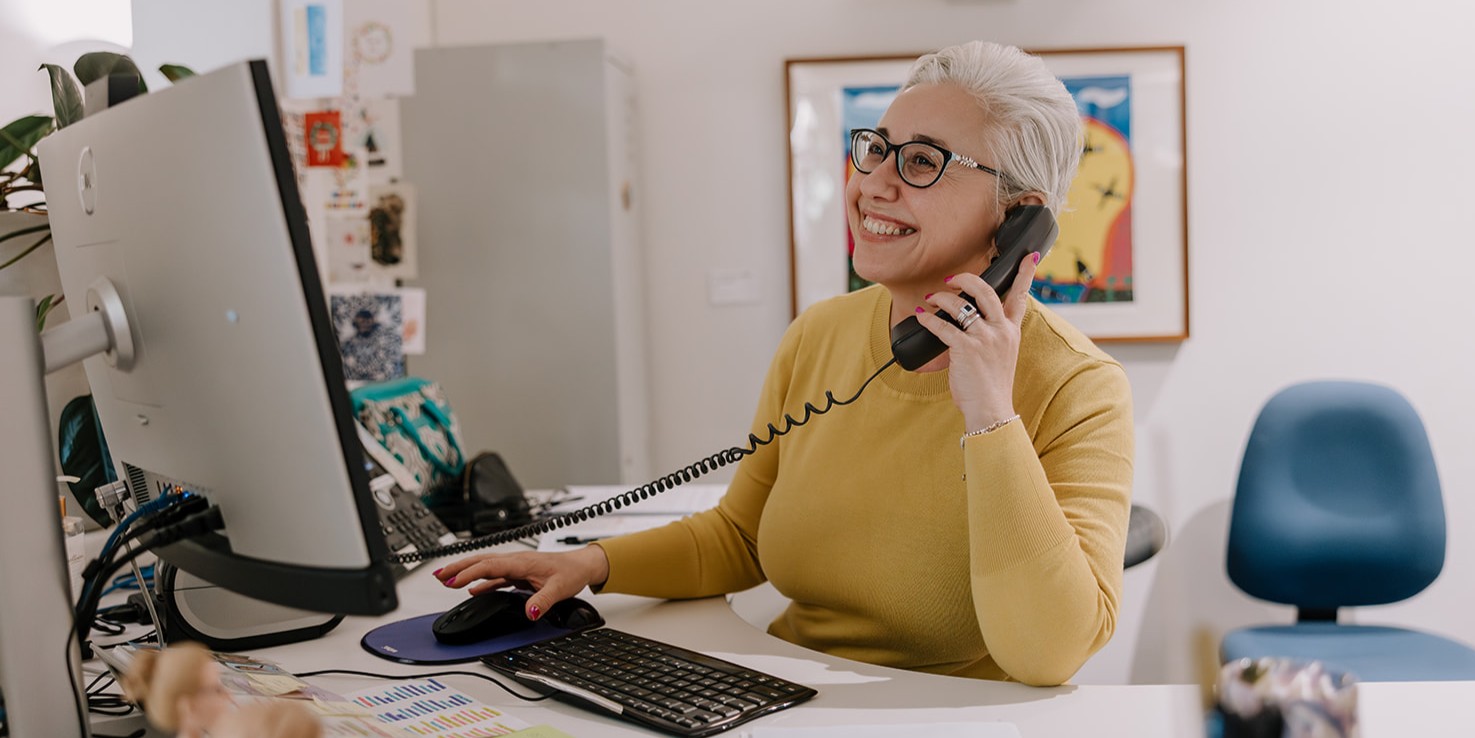 A woman with short, blonde hair and glasses smiles as she talks on a corded black phone at a desk with a computer monitor and keyboard.