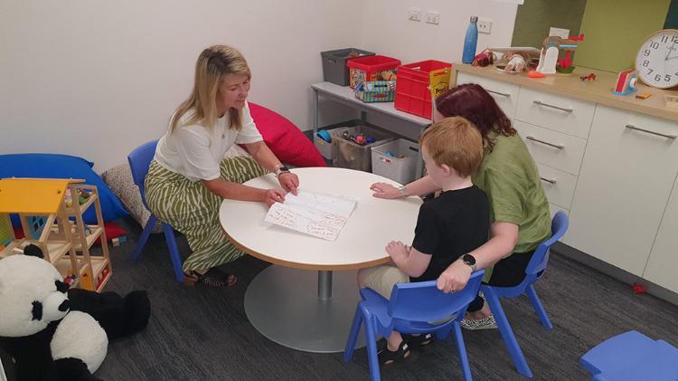 Child sitting with two ladies at a short white round table looking at a piece of paper.