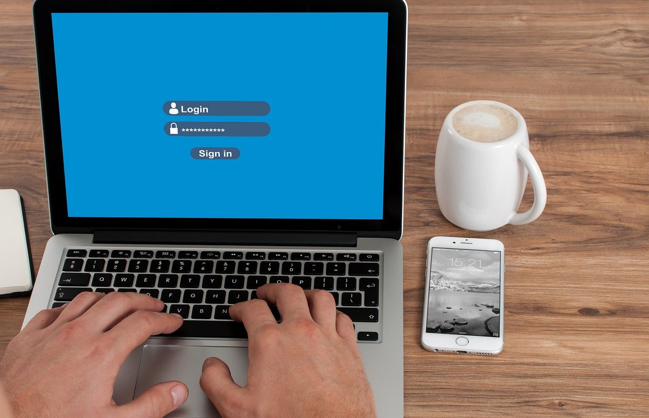 Hands typing on a laptop showing a login form with username and password fields on a blue screen, with a coffee mug and smartphone on the desk nearby.