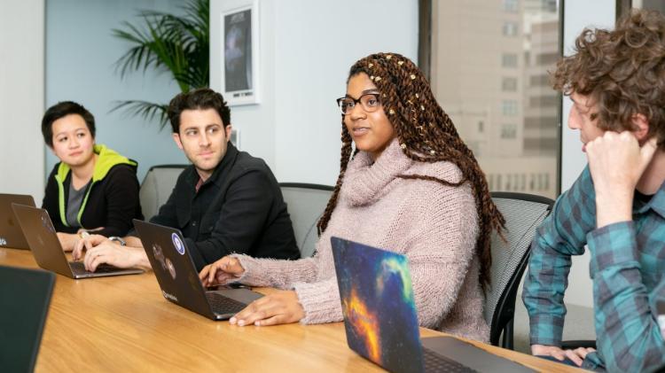 Four people sitting at large table with laptops, focusing to one member talking.