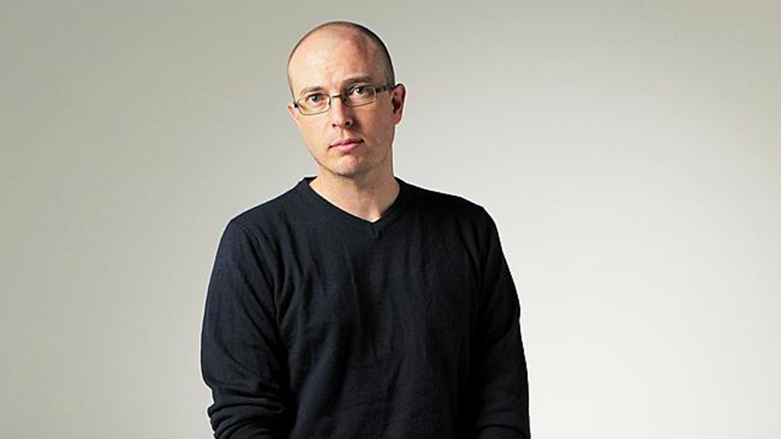 A man with glasses and a black jumper is standing in front of a white backdrop