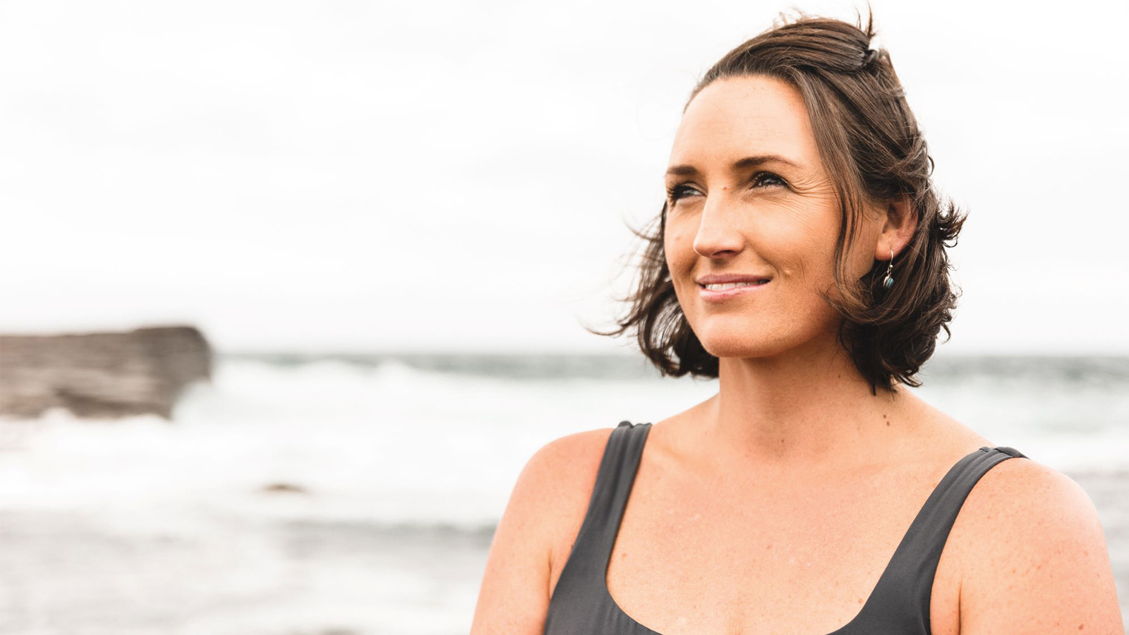 woman with short brown hair is standing in front of the ocean on an overcast day